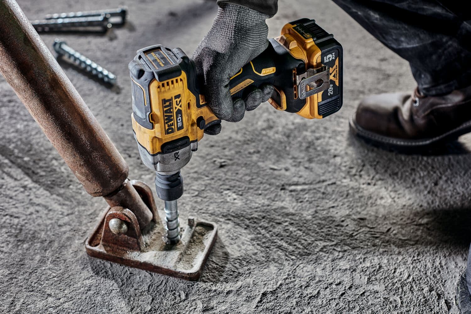 Una persona que lleva guantes de trabajo utiliza una llave de impacto inalámbrica DEWALT 20V (modelo DCF891B) para fijar un perno en una base de metal sobre un suelo de concreto.