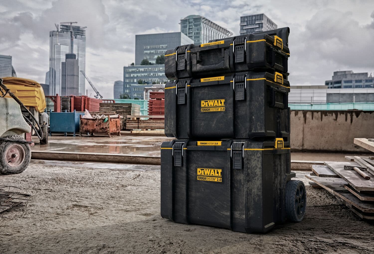 Tres cajas de almacenamiento de herramientas DEWALT ToughSystem 2.0 apiladas con asas amarillas se muestran en un sitio de construcción con edificios modernos al fondo. La caja de almacenamiento inferior tiene ruedas para facilitar el transporte.
