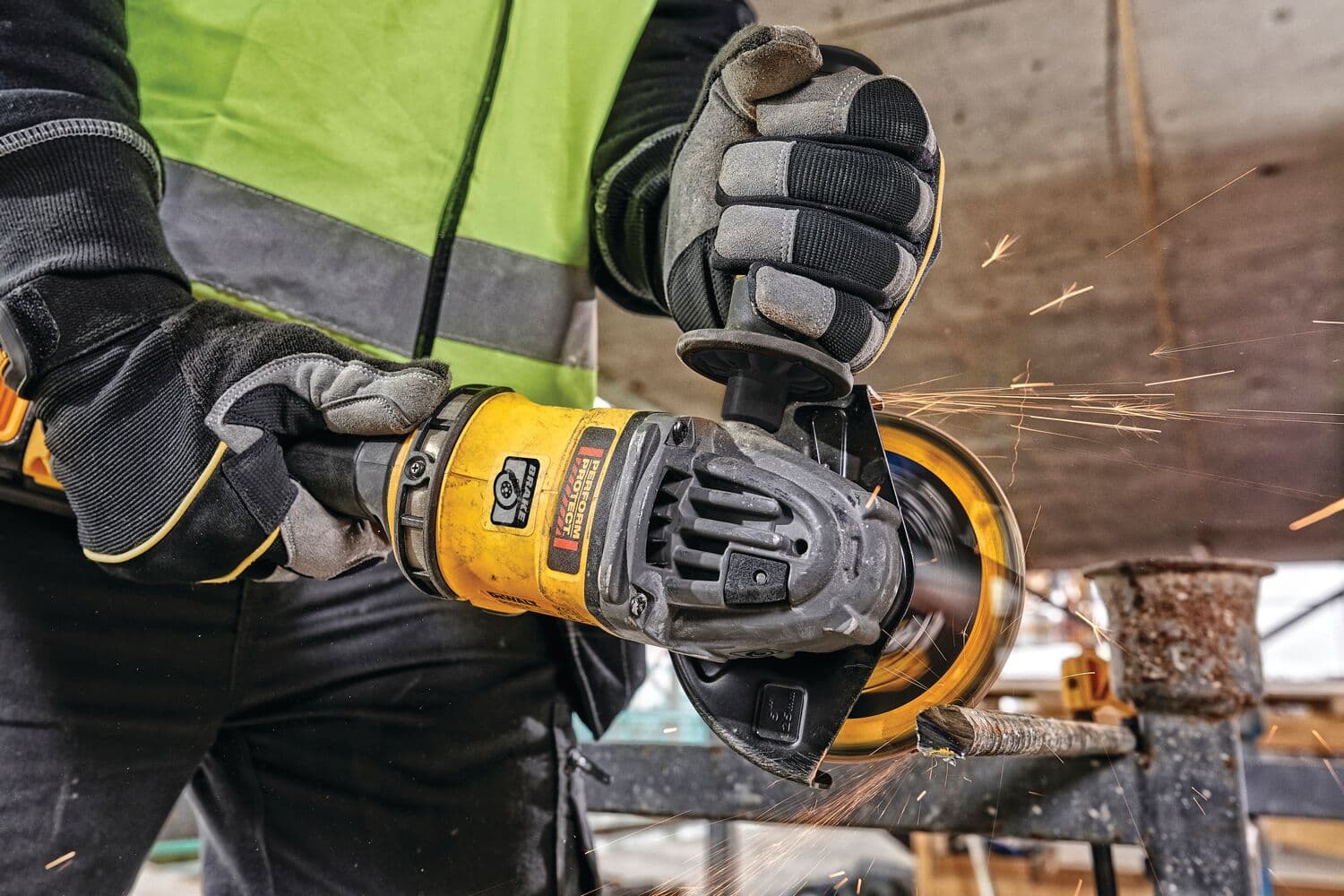 Una persona con guantes de protección y chaleco reflectante utiliza una amoladora angular DEWALT amarilla para cortar metal, generando chispas en un entorno de construcción.