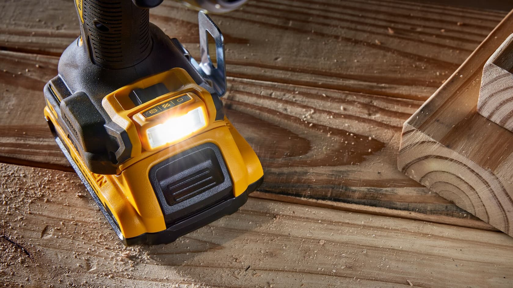 Primer plano de la base de un taladro inalámbrico DEWALT, mostrando la luz de trabajo LED incorporada encendida mientras descansa sobre una superficie de madera con aserrín.