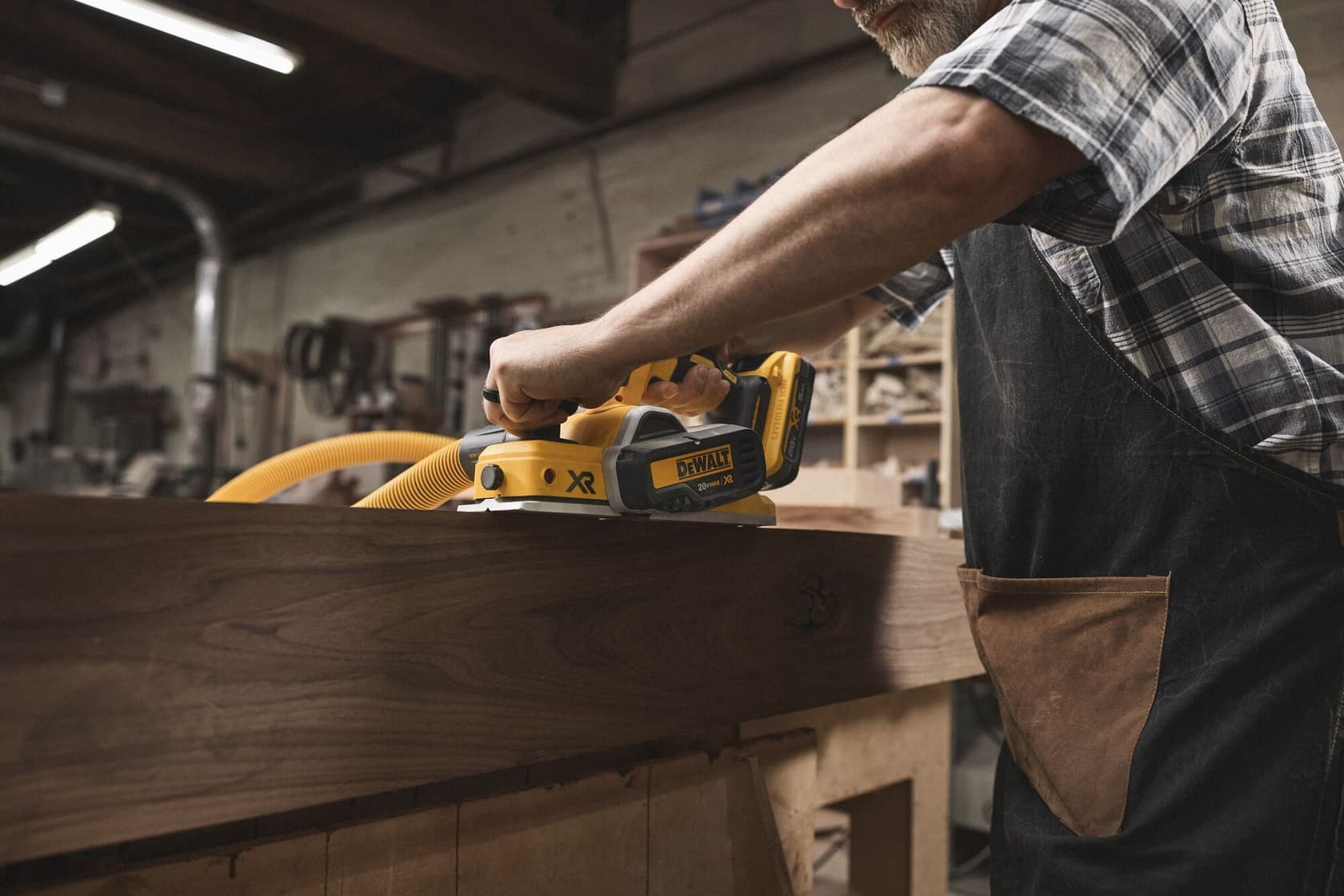 Una persona usa una sierra de carril inalámbrica DEWALT (modelo DCS520B) para cortar una pieza de madera en un taller de carpintería. La persona lleva un delantal y una camisa de cuadros.