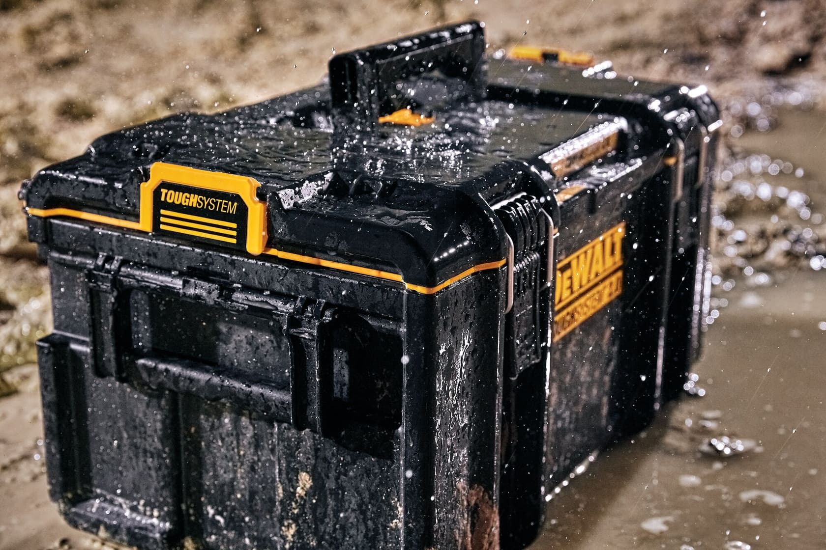 Una caja de almacenamiento DEWALT ToughSystem negra con detalles amarillos, mostrada mojada y embarrada en un sitio de construcción, demostrando su durabilidad y resistencia a la intemperie.