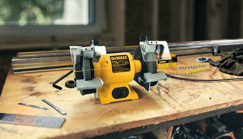 Una esmeriladora de banco DEWALT (modelo DW758) está colocada sobre un banco de trabajo de madera. La herramienta eléctrica amarilla y negra tiene dos ruedas abrasivas, una a cada lado, con protectores y apoyos. Alrededor de la esmeriladora hay limas metálicas, varillas y pequeñas herramientas de taller en un entorno de taller.